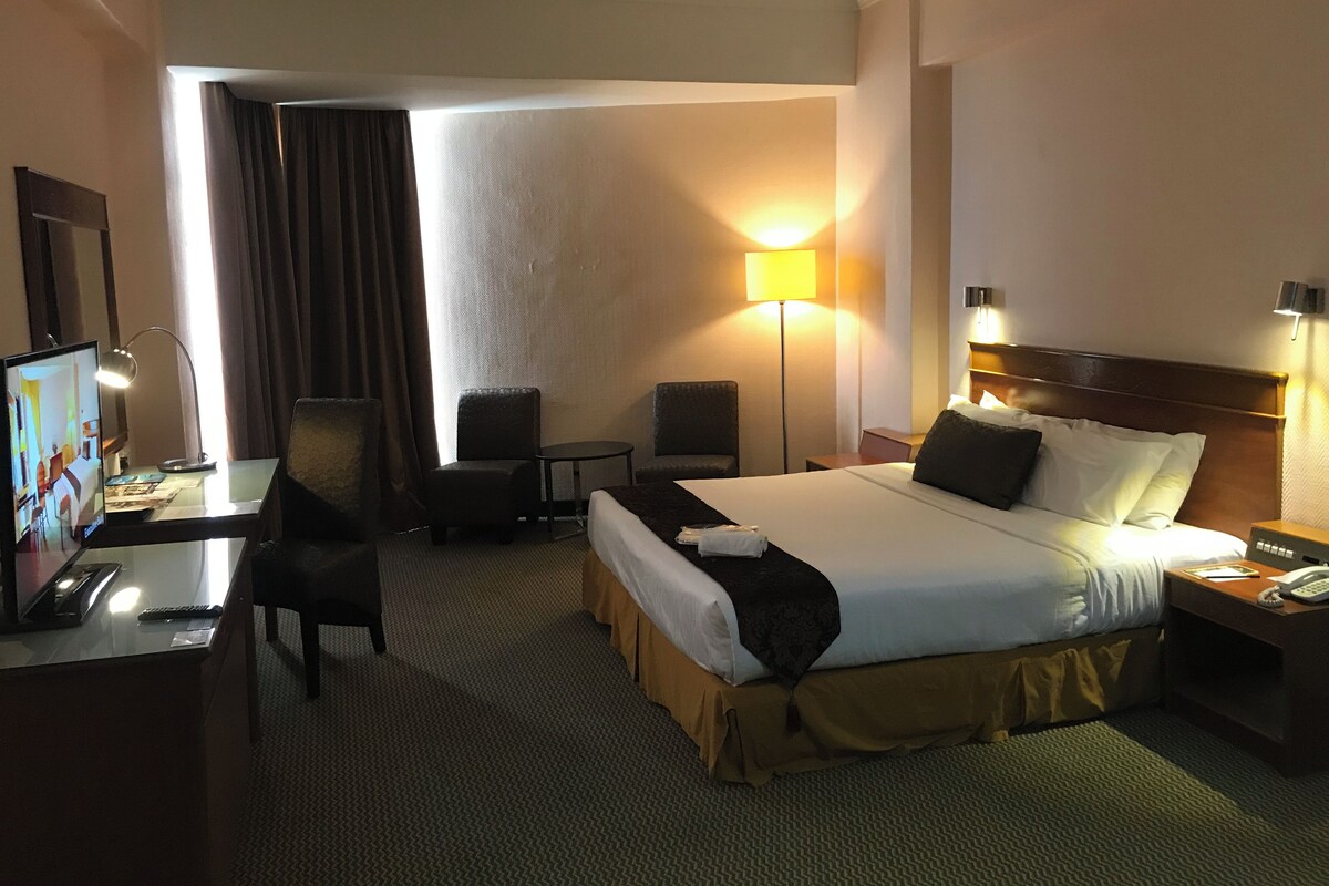 Habitación de hotel con cama, escritorio con computadora, dos sillas, lámpara y televisor.