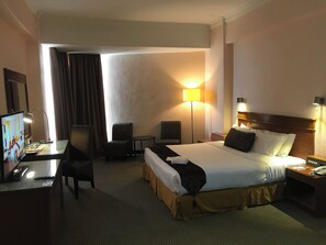 A hotel room with a bed, a desk with a computer, two chairs, a lamp, and a TV.