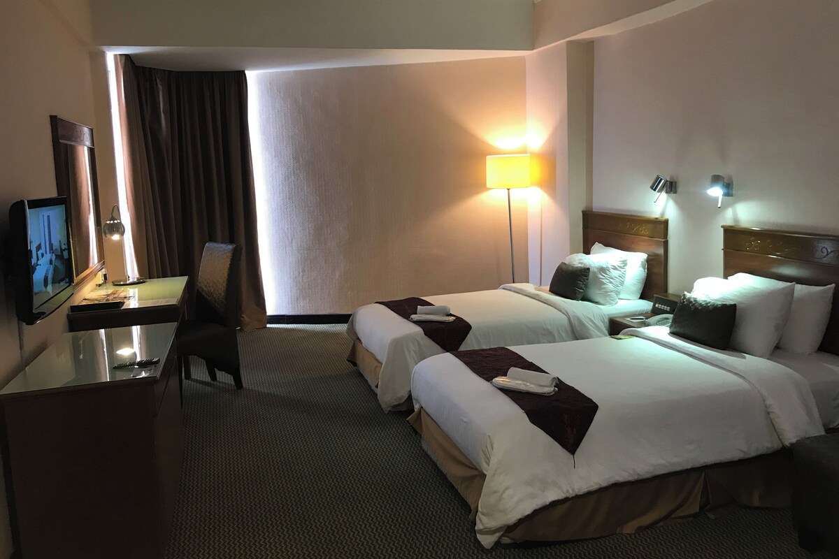 A hotel room with two beds, a desk with a television, a lamp, and a view of the outside.