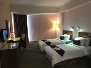 Habitación de hotel con dos camas, un escritorio con televisor, una lámpara y vistas al exterior.