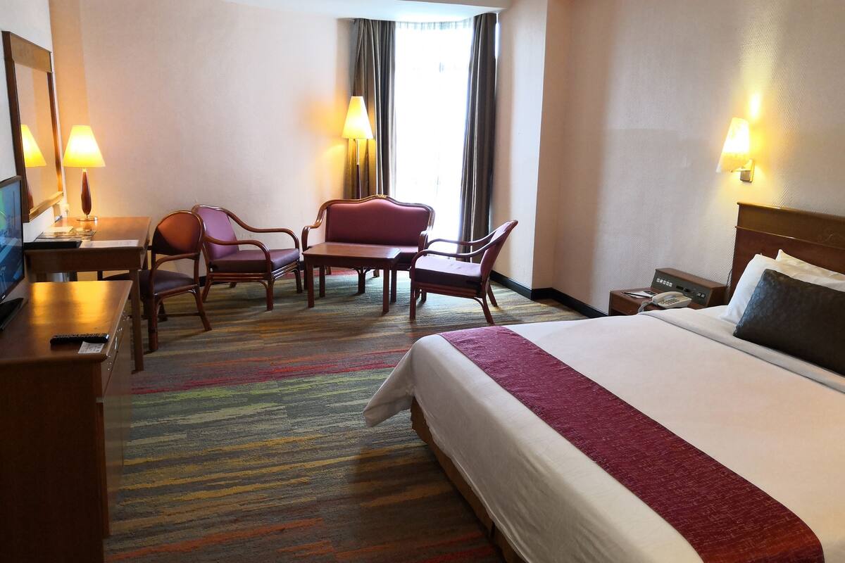 Une chambre d’hôtel comprenant un lit, un bureau avec un ordinateur, deux chaises, une petite table et une fenêtre avec des rideaux.