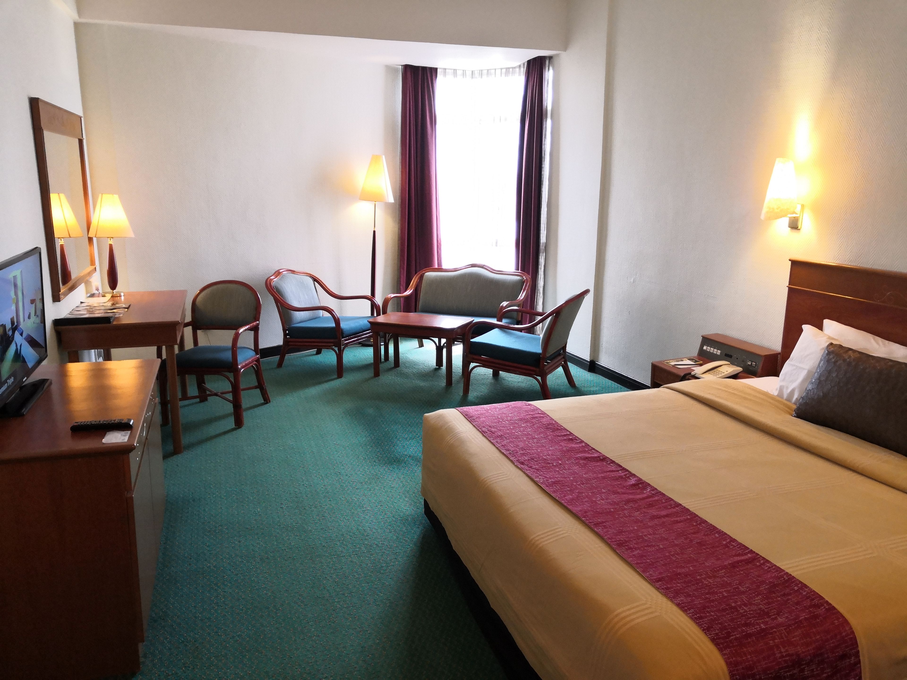 Quarto de hotel com cama, duas poltronas, uma mesinha, escrivaninha com computador e televisão.