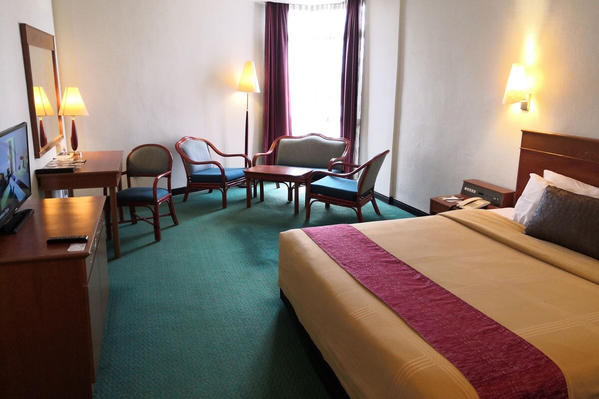 Una habitación de hotel con una cama, dos sillones, una mesita, un escritorio con una computadora y un televisor.