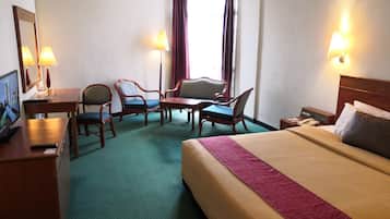 Quarto de hotel com cama, duas poltronas, uma mesinha, escrivaninha com computador e televisão.