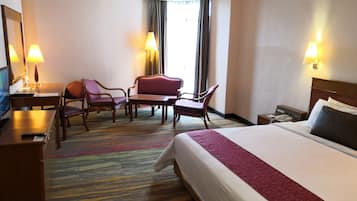 A hotel room with a bed, a desk with a computer, two chairs, a small table, and a window with curtains.