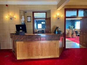 Un comptoir d'accueil avec un ordinateur portable, un téléphone et une petite table sur laquelle est posée une plante. Au premier plan, on aperçoit un espace recouvert d'un tapis rouge, et au fond, une salle à manger avec des chaises et une table.