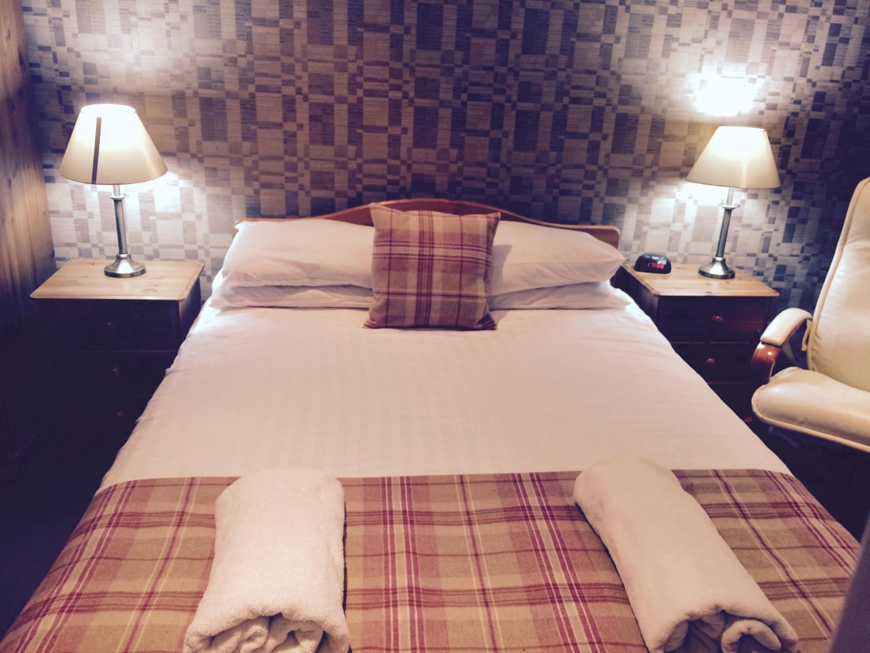 Una habitación de hotel con una cama, dos mesitas de noche con lámparas, una silla y una manta a cuadros.