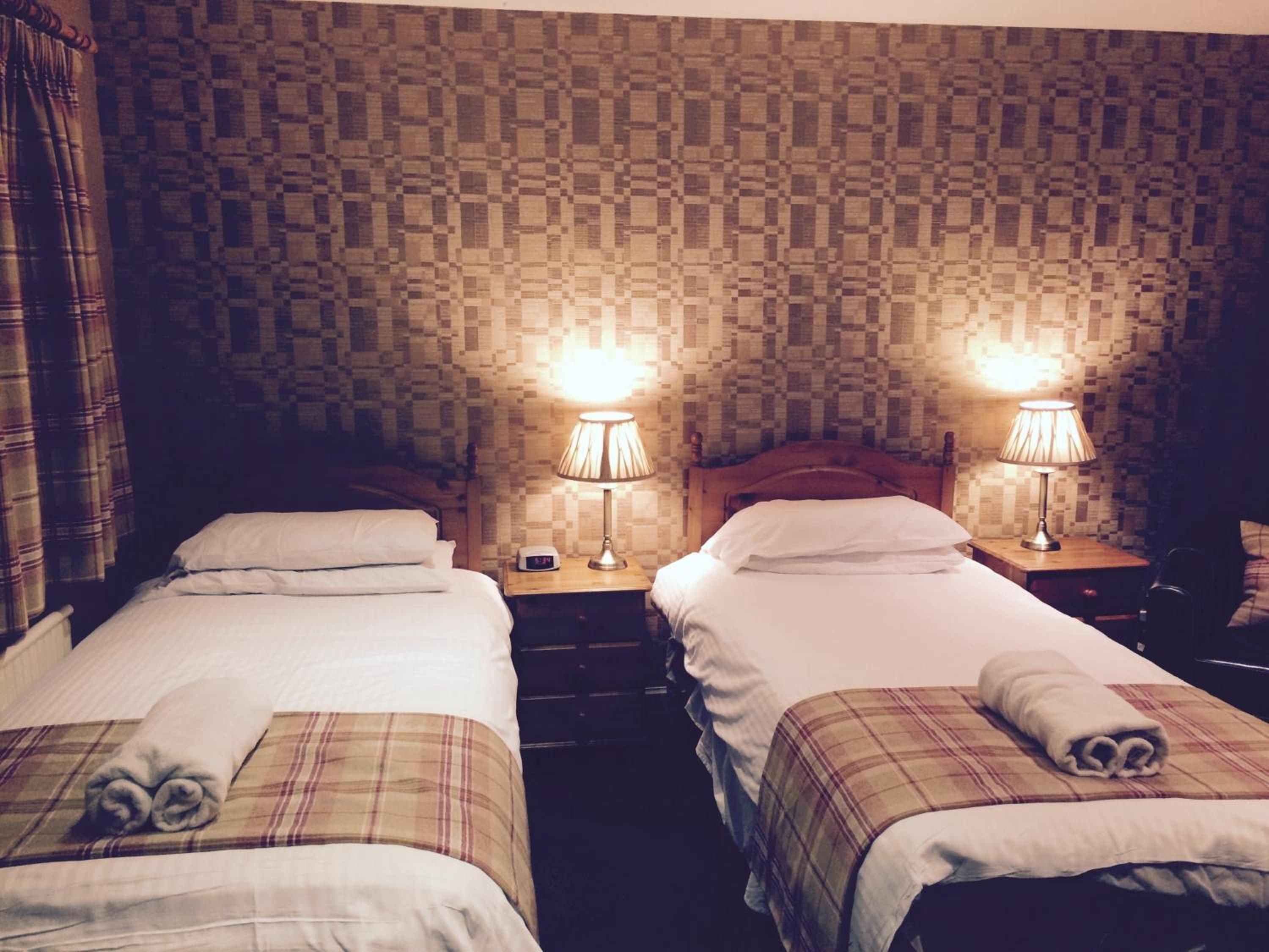 Habitación de hotel con dos camas, cada una con almohada y toalla enrollada. Hay dos mesitas de noche con lámparas y un reloj. Las paredes tienen un diseño de cuadros.
