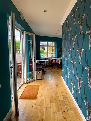 Un pasillo con piso de madera, un comedor con mesas y sillas, y una pared verde azulado con motivos de ciervos.