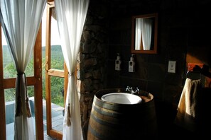A rustic bathroom with a stone wall, wooden barrel sink, and a view of greenery outside. - Eagle Tented Lodge (Okaukuejo)