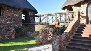 A stone building with a thatched roof, a staircase leading to a wooden deck, and a small garden area with potted plants. - Eagle Tented Lodge (Okaukuejo)
