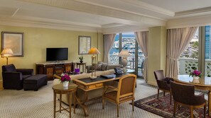 A hotel room with a sofa, armchairs, a desk, a television, and a view of the marina.