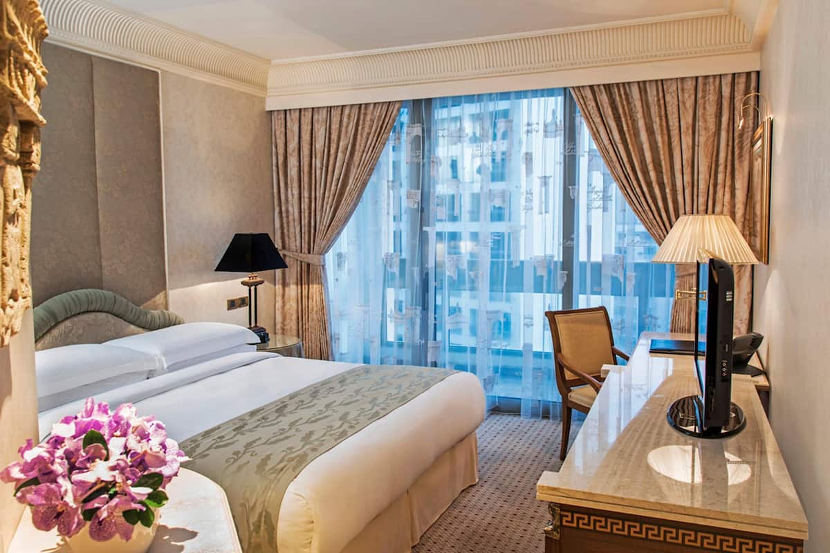 Habitació d'hotel amb un llit gran, escriptori amb làmpada, una cadira i vista a un edifici a través de cortines translúcides.