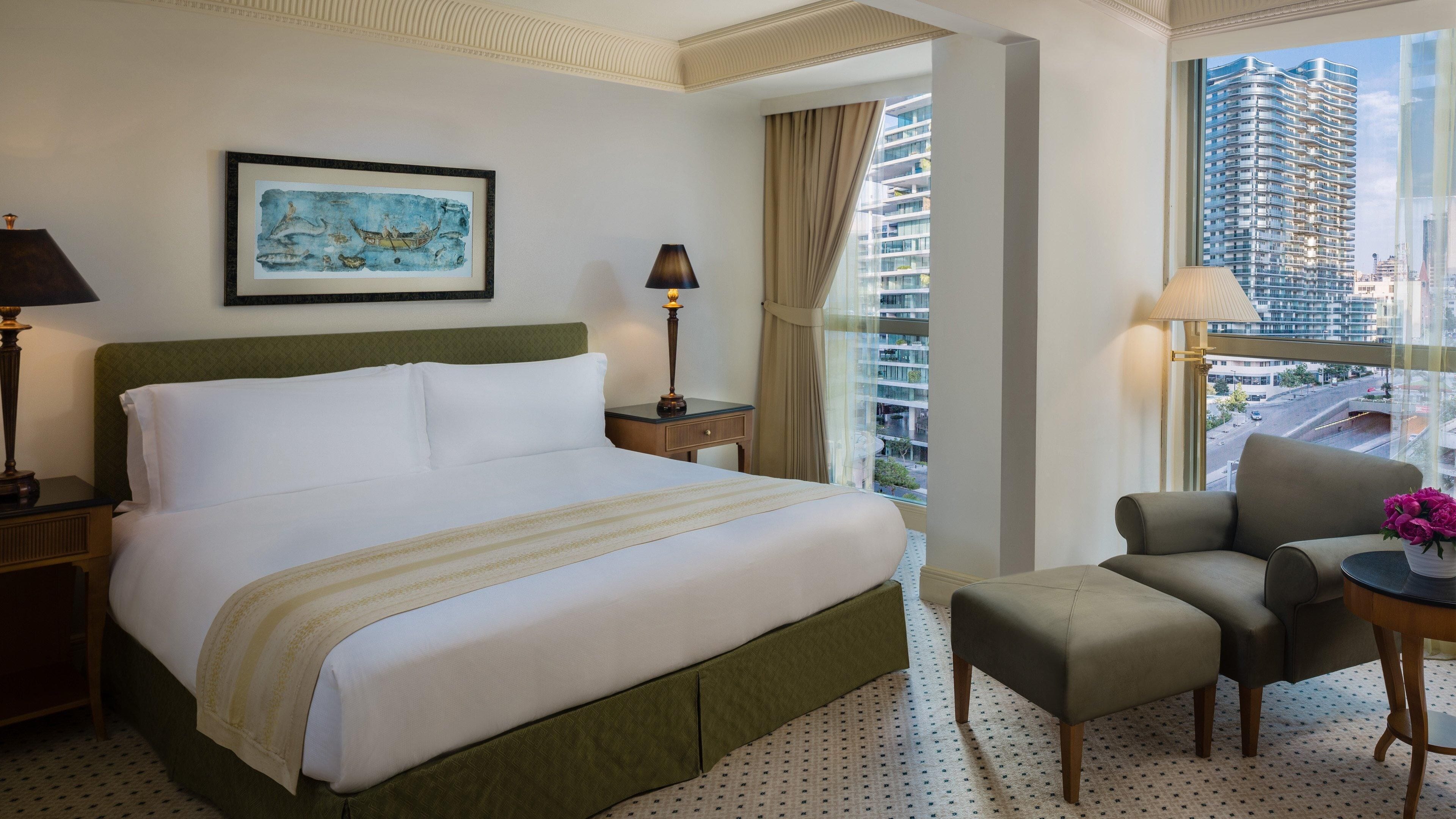 Una camera d'albergo con un letto grande, una sedia, un comodino, una lampada, un quadro incorniciato e vista sulla città.