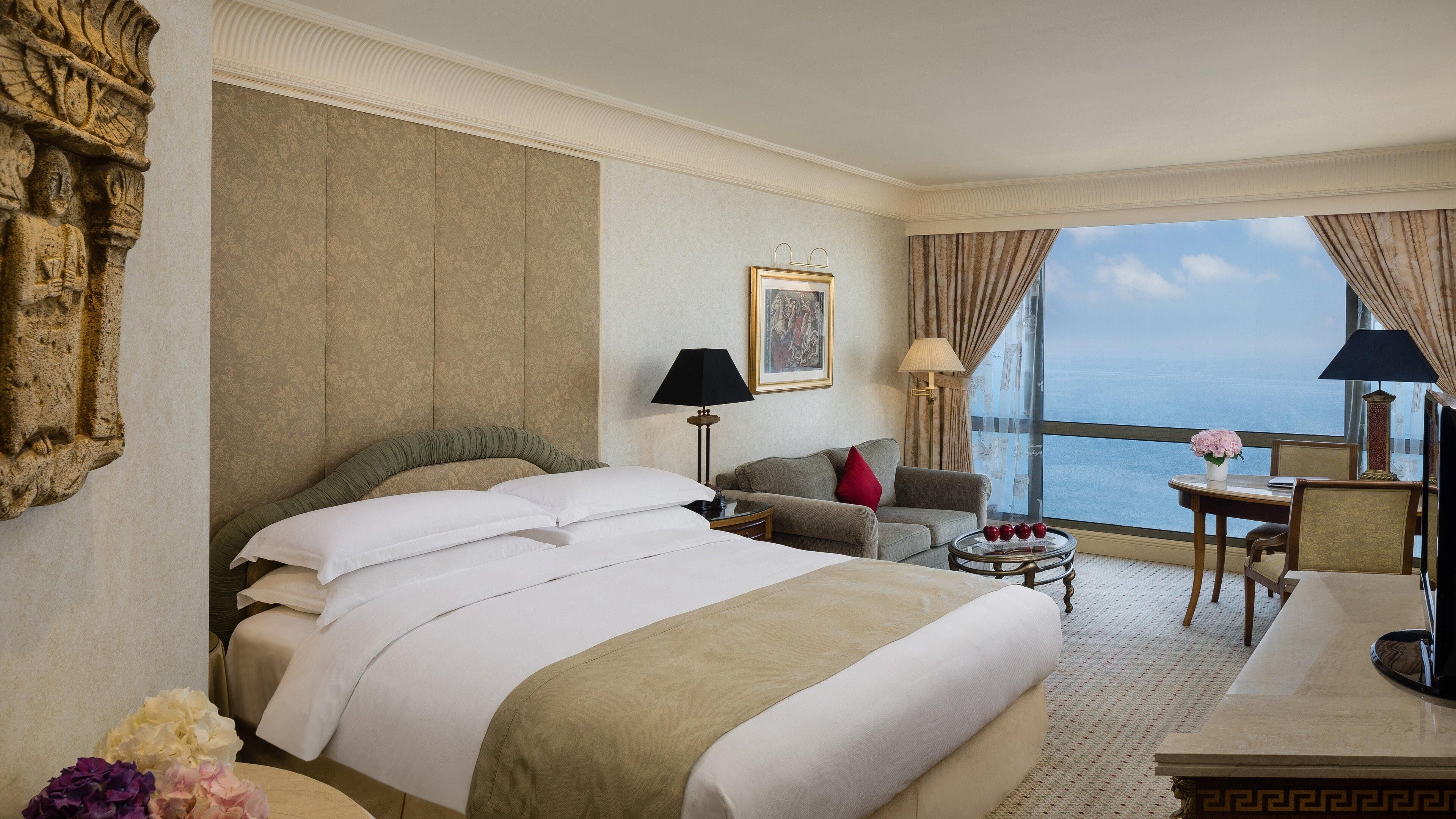 Camera d'albergo con un letto grande, un divano, un tavolino con un vaso di fiori e vista sul mare attraverso ampie finestre.