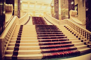 A grand staircase with marble steps and ornate railings, leading to a bright, well-lit area.