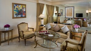A hotel room with a sofa, armchairs, a glass coffee table, a desk with a lamp, a television, and artwork on the walls.