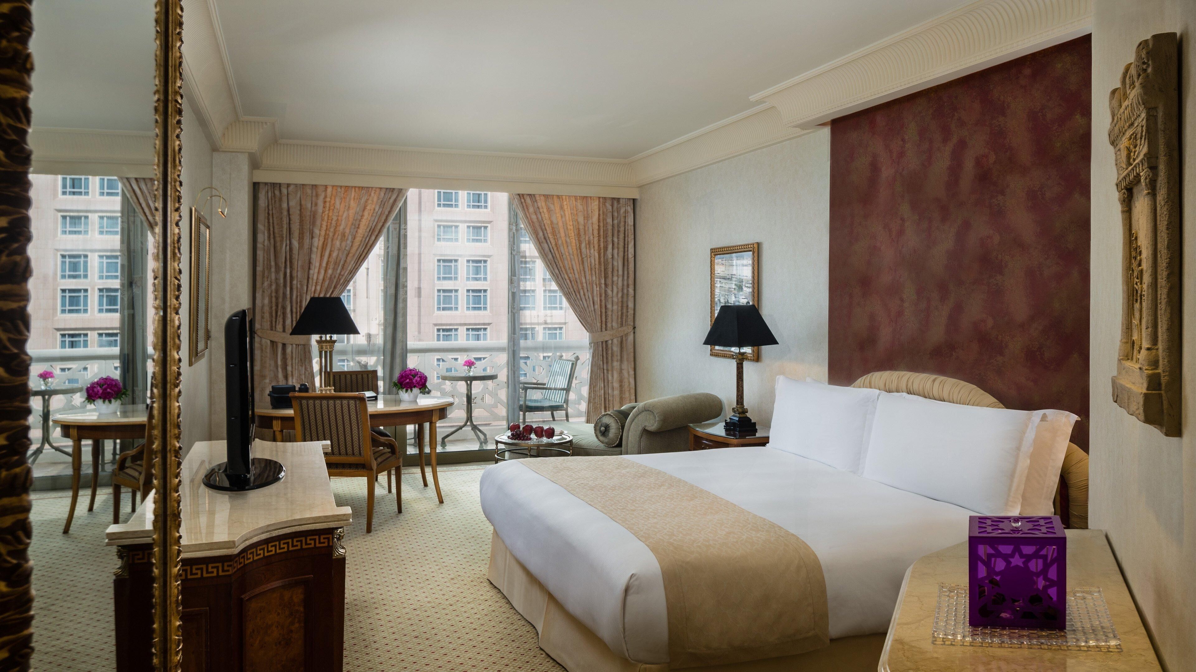 Una camera d'albergo con un letto grande, una scrivania con lampada, una sedia, un tavolino con fiori e vista sugli edifici attraverso ampie finestre.