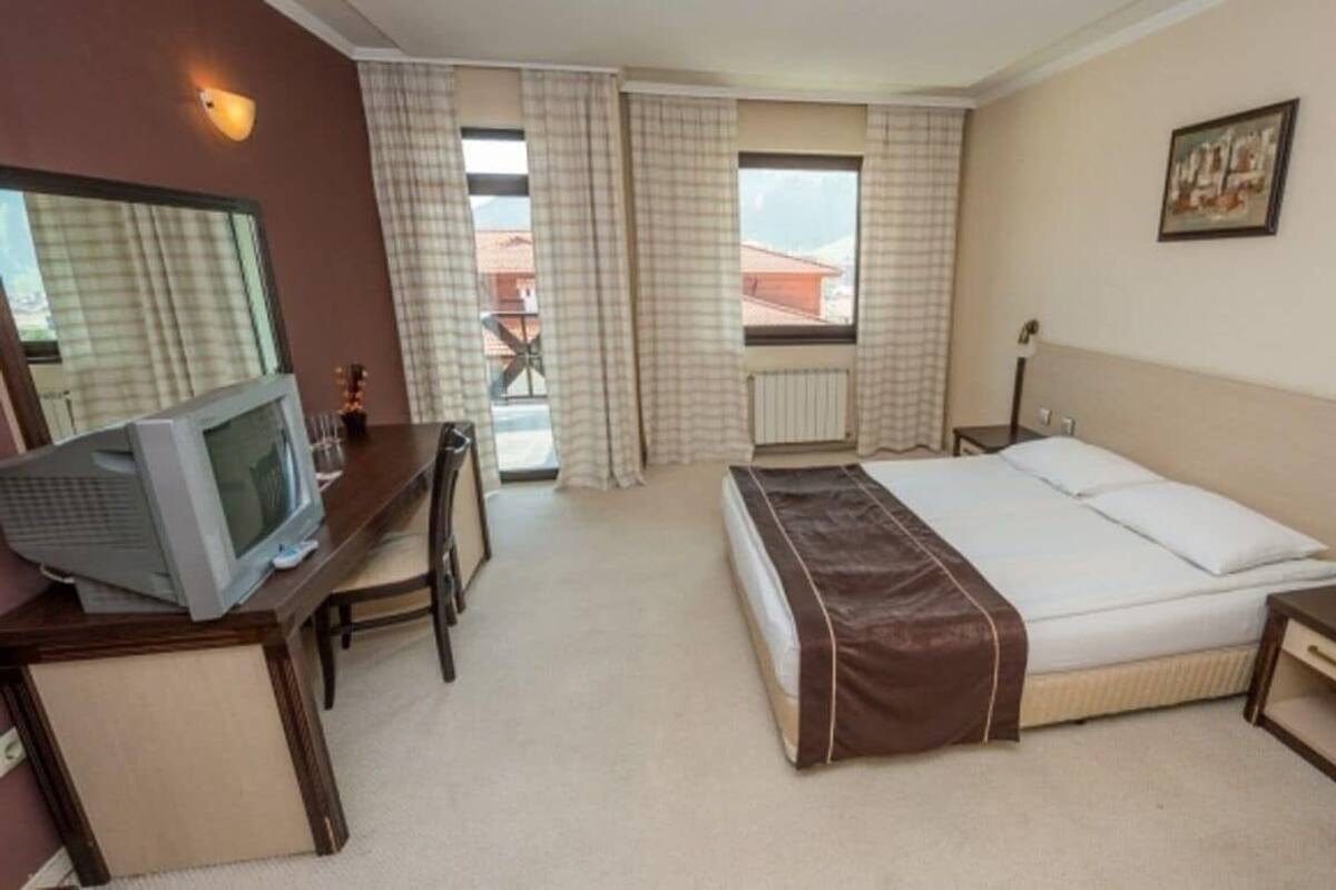 A hotel room with a bed, a desk with a television, a window with curtains, and a radiator.