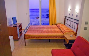 A hotel room with a bed, a desk, a chair, a TV, and a view of the beach.