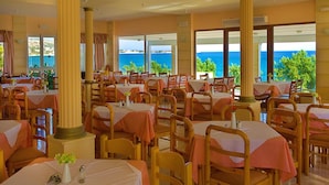 Een restaurant met meerdere tafels en stoelen, grote ramen met uitzicht op zee, en een warme kleurenschema.
