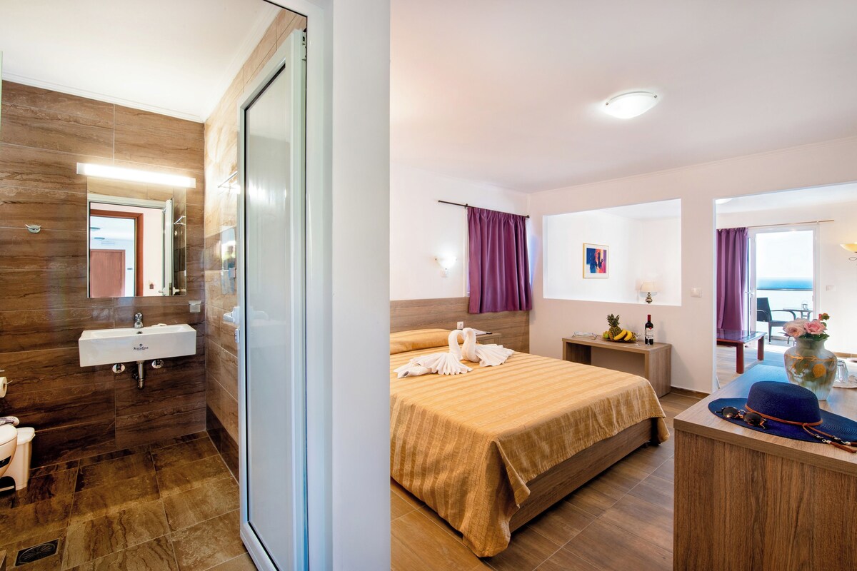 A hotel room with a wooden floor, a bed with a striped bedspread, a bedside table with a lamp, a small table with fruits and a bottle, and a view of the sea through a balcony door.