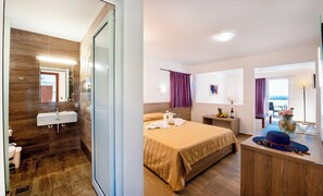 A hotel room with a wooden floor, a bed with a striped bedspread, a bedside table with a lamp, a small table with fruits and a bottle, and a view of the sea through a balcony door. - Ilyssion (Rhodes)