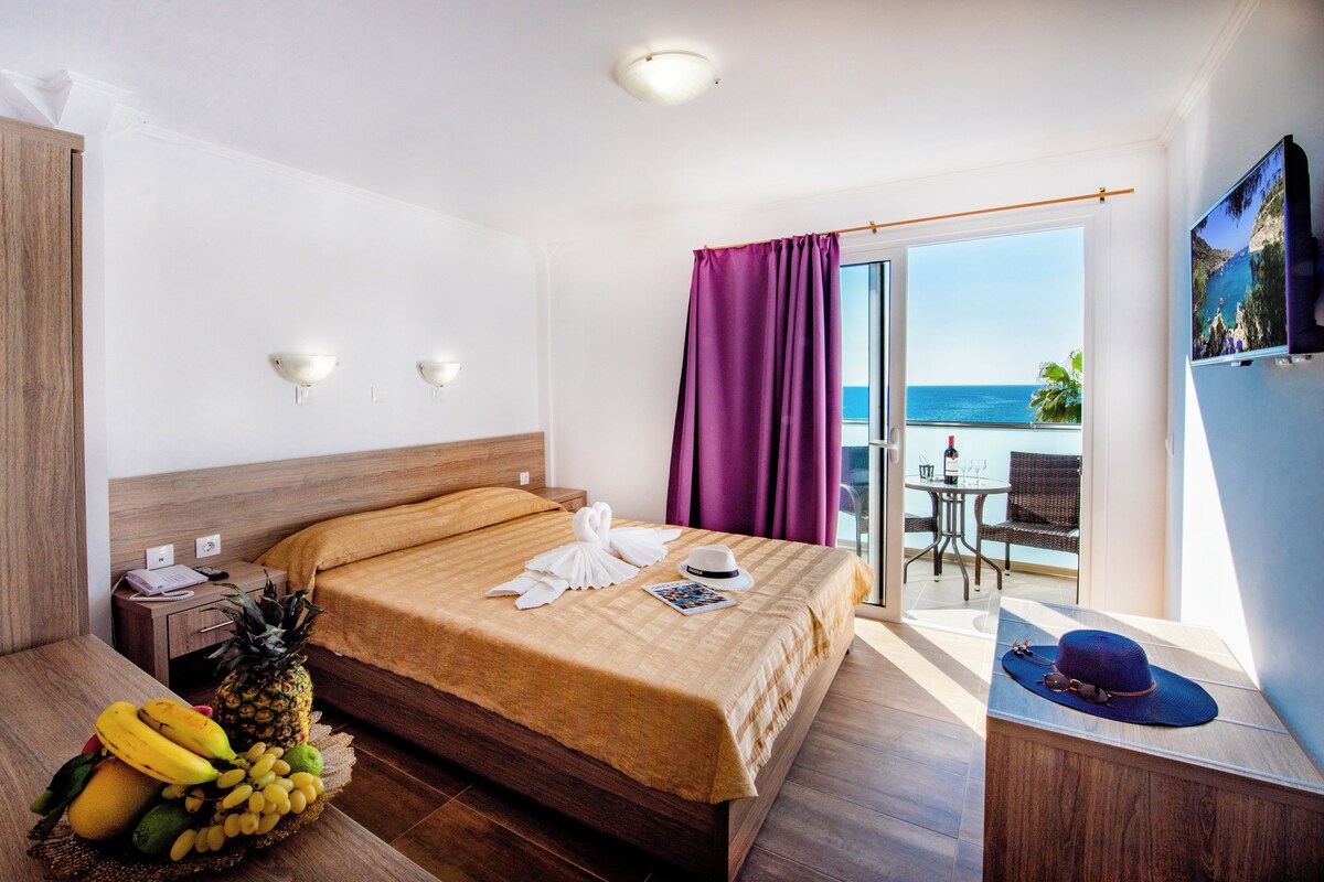Una habitación de hotel con una cama, una cesta de frutas, un sombrero y vistas al mar.