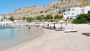 Una playa con tumbonas y sombrillas, una colina rocosa al fondo y edificios con balcones.
