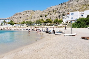 A beach with sun loungers and umbrellas, a rocky hillside in the background, and buildings with balconies.