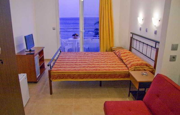 A bedroom with a bed, a desk, a chair, a TV, and a view of the sea.