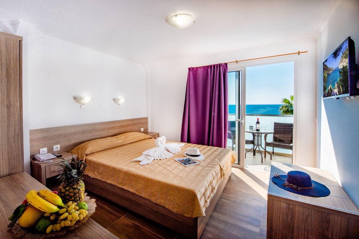 Habitación de hotel con una cama, una cesta de frutas, un sombrero y vistas al océano.