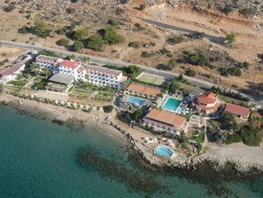 Vista aérea de un complejo turístico costero con varios edificios, una playa y aguas azules cristalinas.