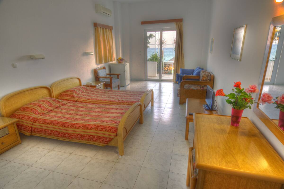 Habitación de hotel con dos camas, un escritorio, una silla, una mesita, una planta en maceta y un ventanal corredero que da a un balcón con vista al océano.
