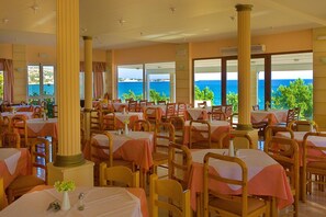 Un restaurante con varias mesas y sillas, grandes ventanales con vista al mar y una decoración en tonos cálidos.