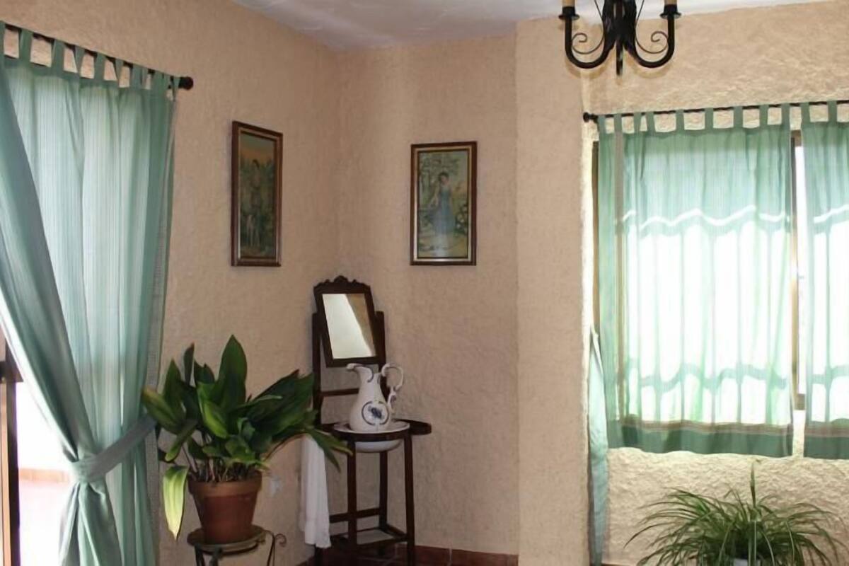 A room with a large window, a potted plant, a small table with a mirror, and two framed pictures on the wall.