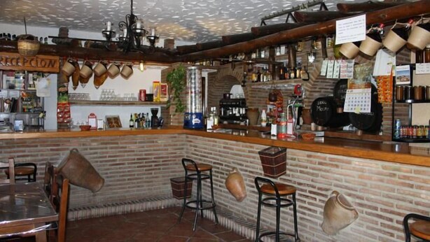Un espace bar avec un mur en briques, un comptoir en bois et des tabourets. On y trouve des chopes suspendues, un calendrier, ainsi que diverses bouteilles et verres sur les étagères.