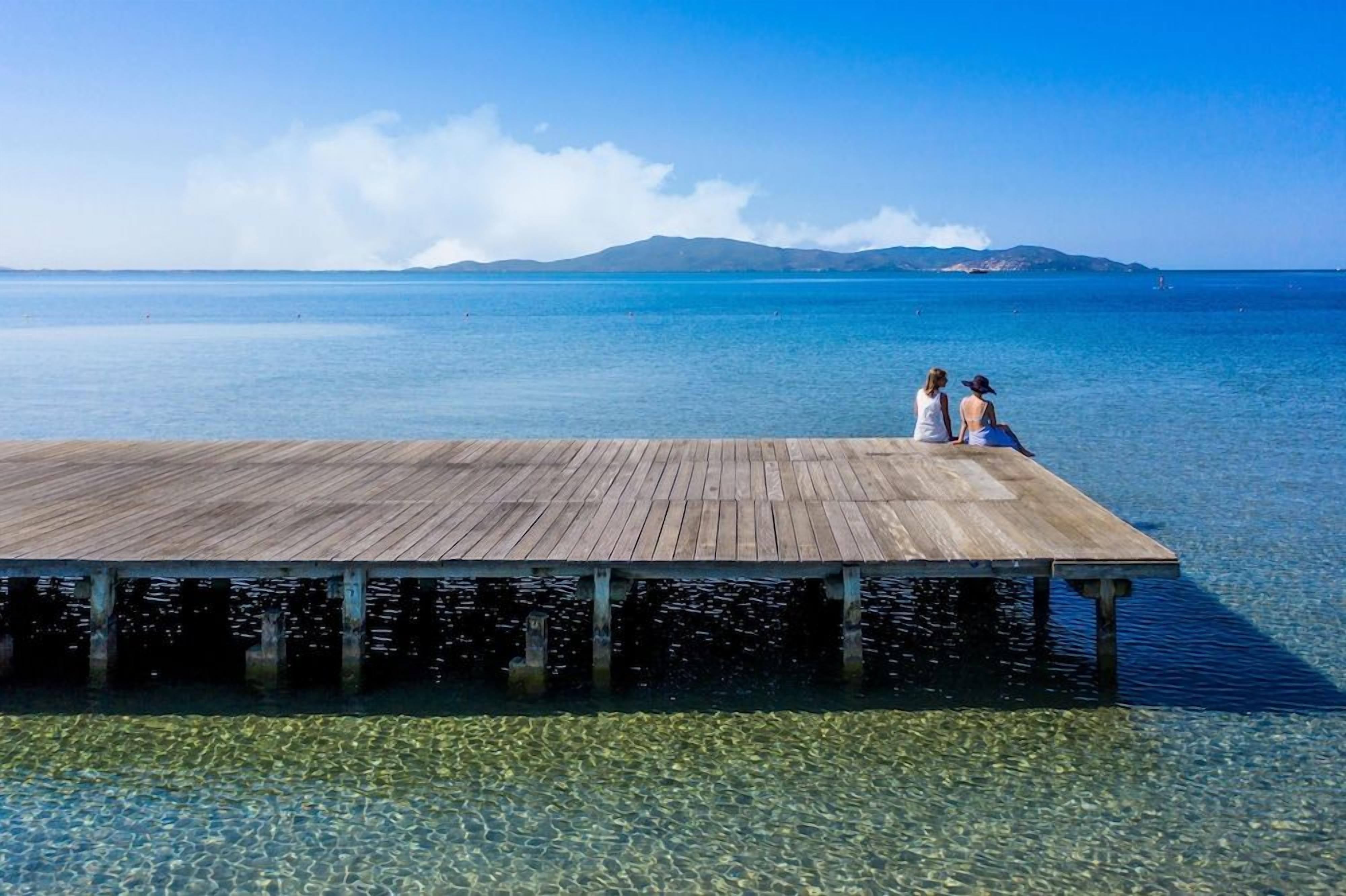 A wooden pier extending into clear blue water with two people sitting at the end.