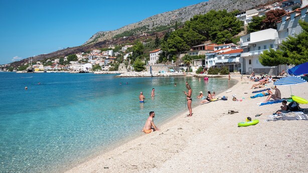 Une plage côtière aux eaux cristallines, où l’on peut voir des gens se baigner et se détendre sur le sable, ainsi que des bâtiments qui s’étendent le long du flanc de la colline.