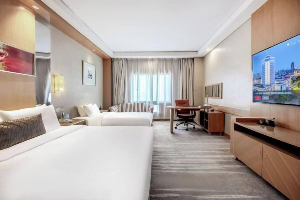 A hotel room with two beds, a desk, a chair, a TV, and a cityscape view.