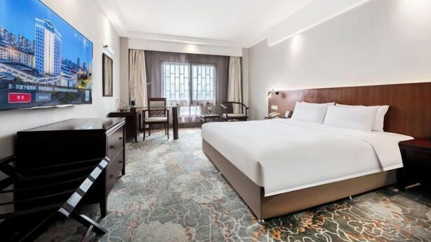 Ein Hotelzimmer mit einem großen Bett, einem Schreibtisch, einem Sessel, einem an der Wand befestigten Fernseher und einem Fenster mit Vorhängen.