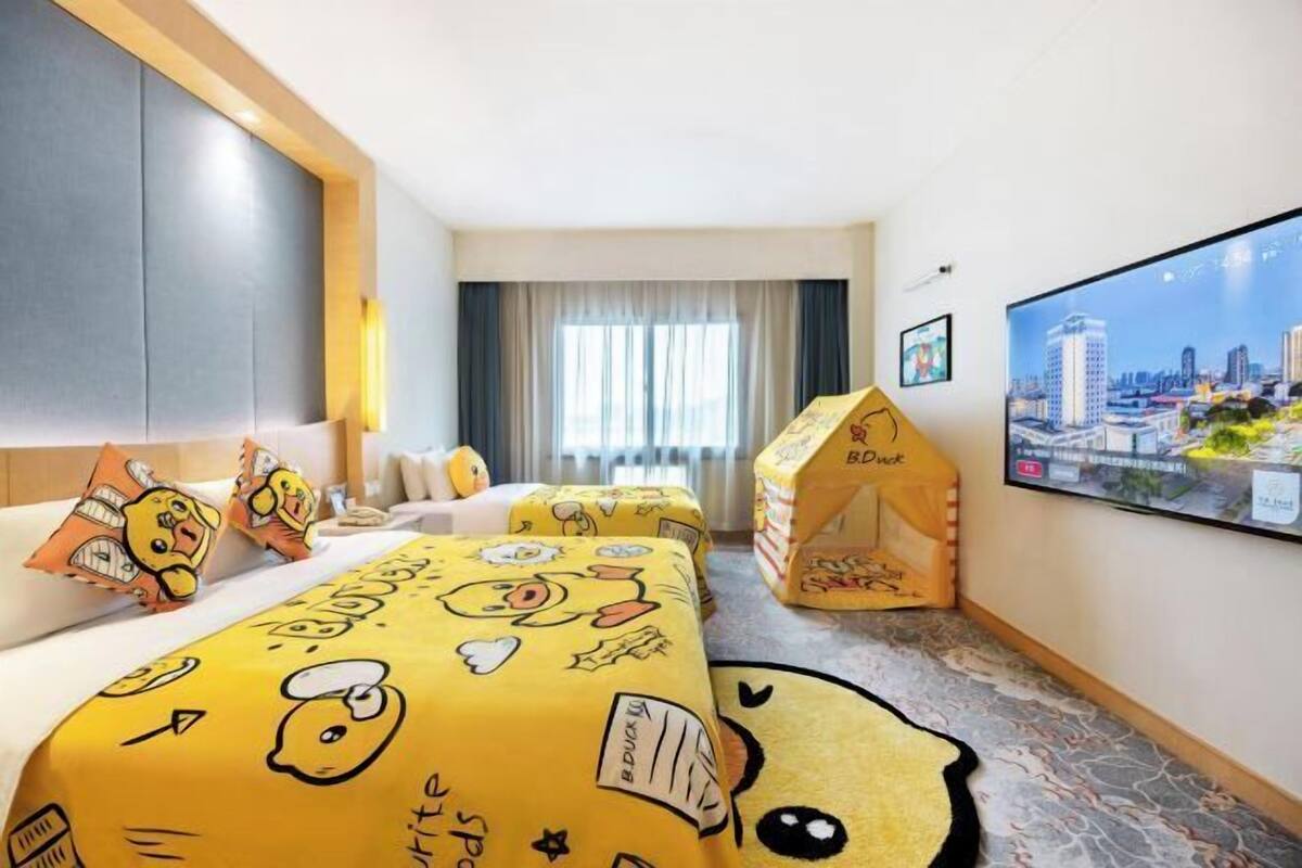 A hotel room with a bed covered in yellow Duck-themed bedding, a cityscape view on the wall, and a small playhouse in the corner.