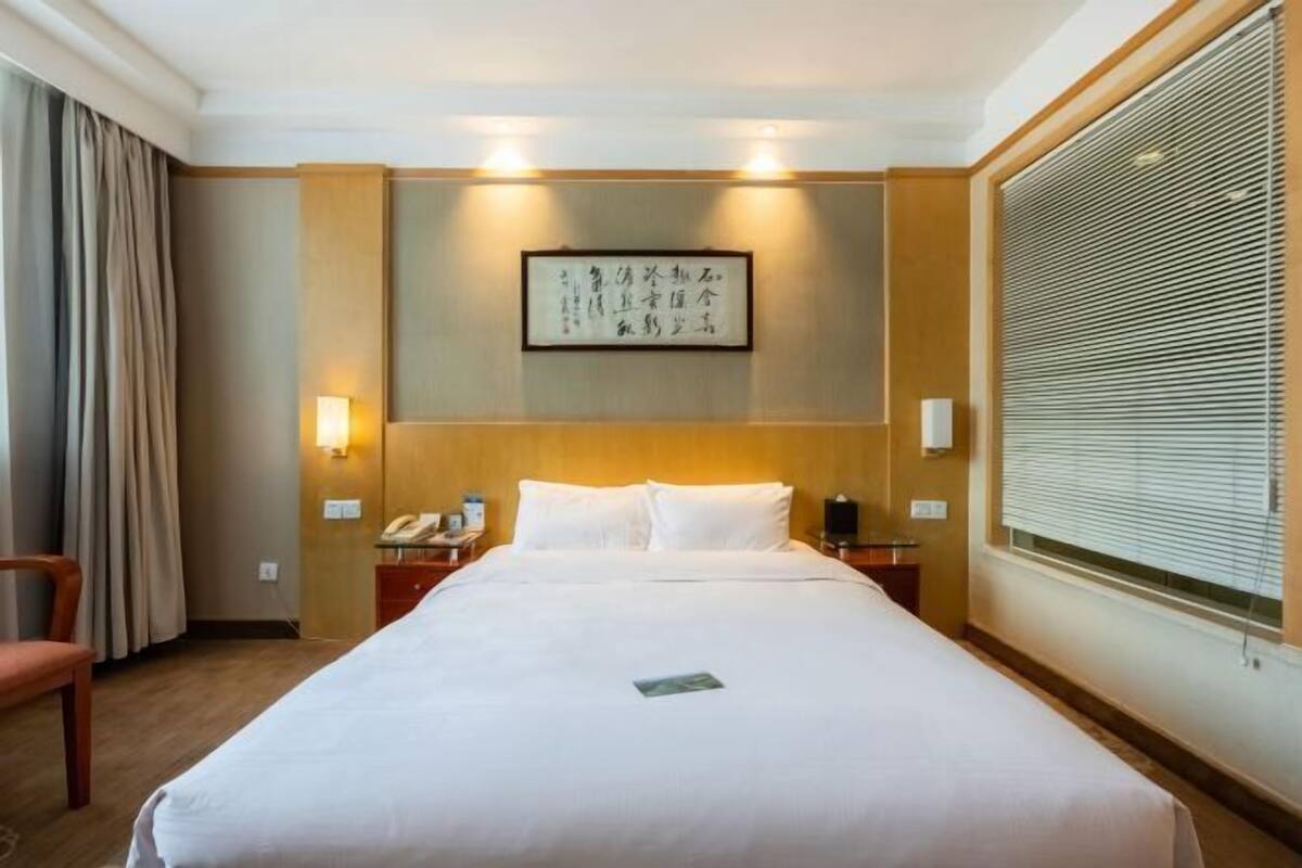 A hotel room with a large bed, a bedside table with a lamp, a telephone, and a window with blinds.