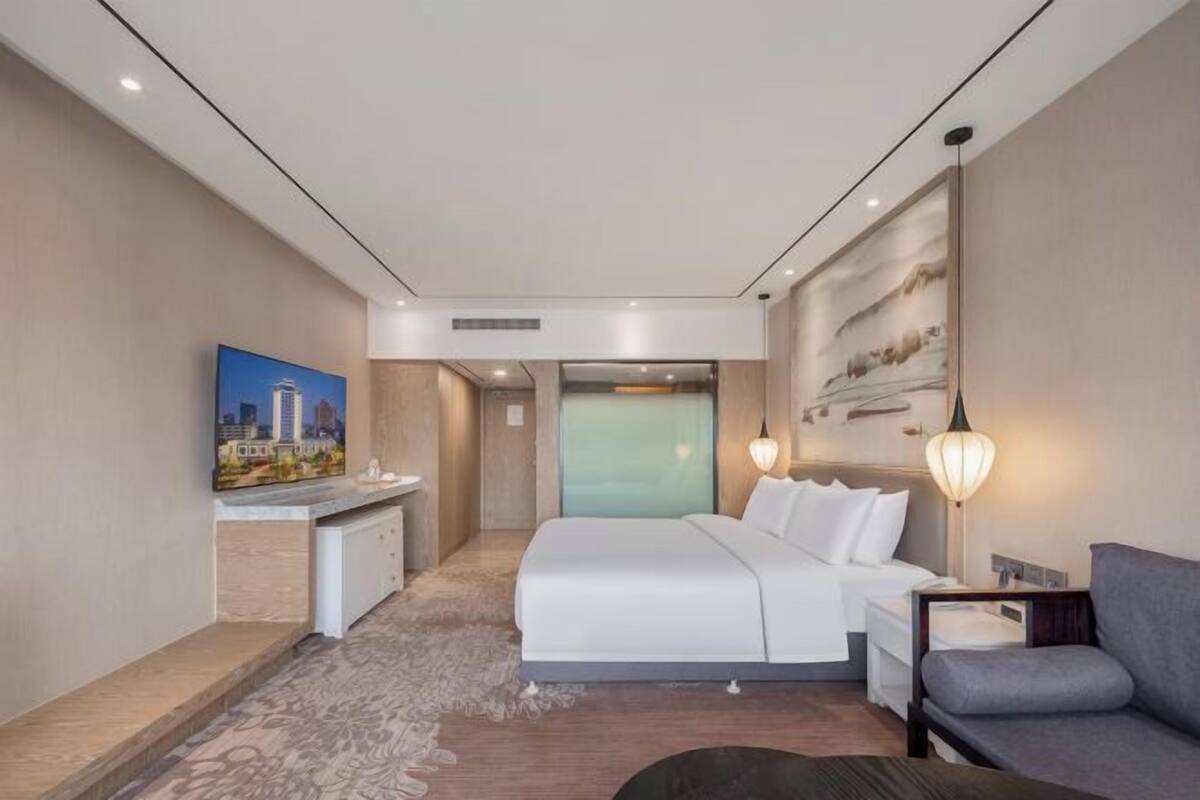 A modern hotel room with a large bed, a desk, a sofa, and a painting on the wall.