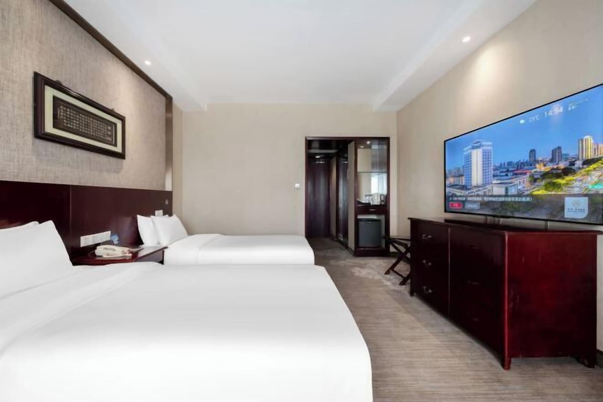 A hotel room with two beds, a TV displaying a cityscape, and a wooden dresser.