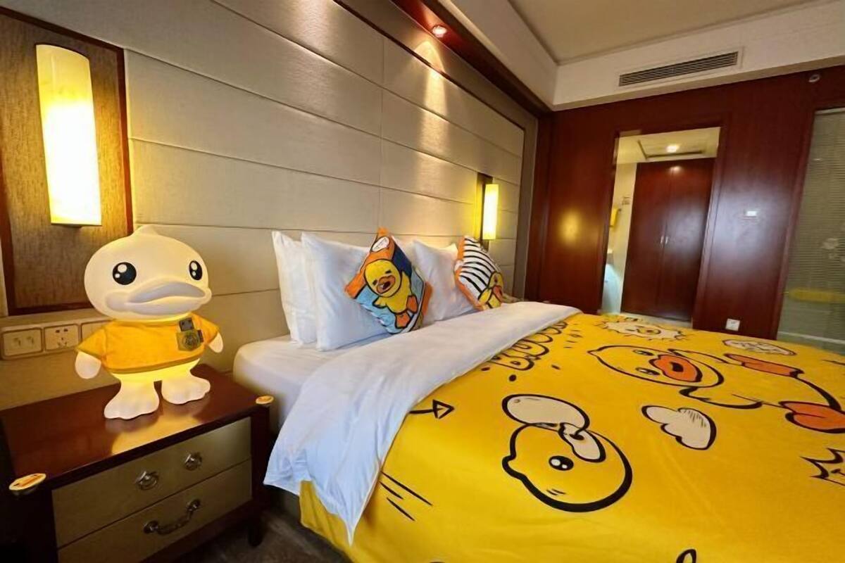 A hotel room with a bed featuring cartoon character bedding, a nightstand with a duck-shaped night light, and a wooden headboard.