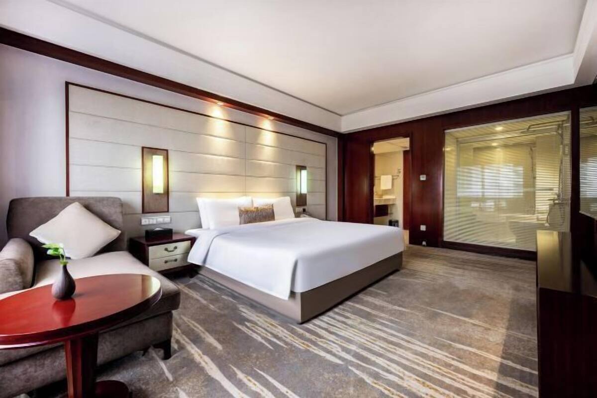 A modern hotel room with a large bed, a sofa, a small table, and a large window with blinds.