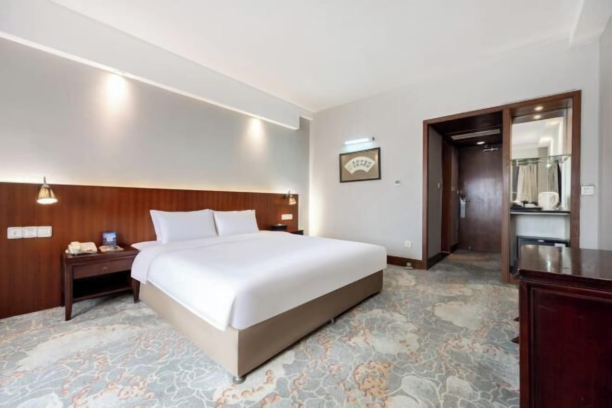 A hotel room with a large bed, bedside tables, a wooden headboard, a carpeted floor, and a door leading to another room.