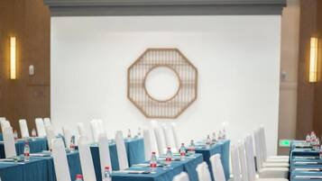 Un salón de conferencias con filas de mesas, sillas blancas y un elemento decorativo en la pared.