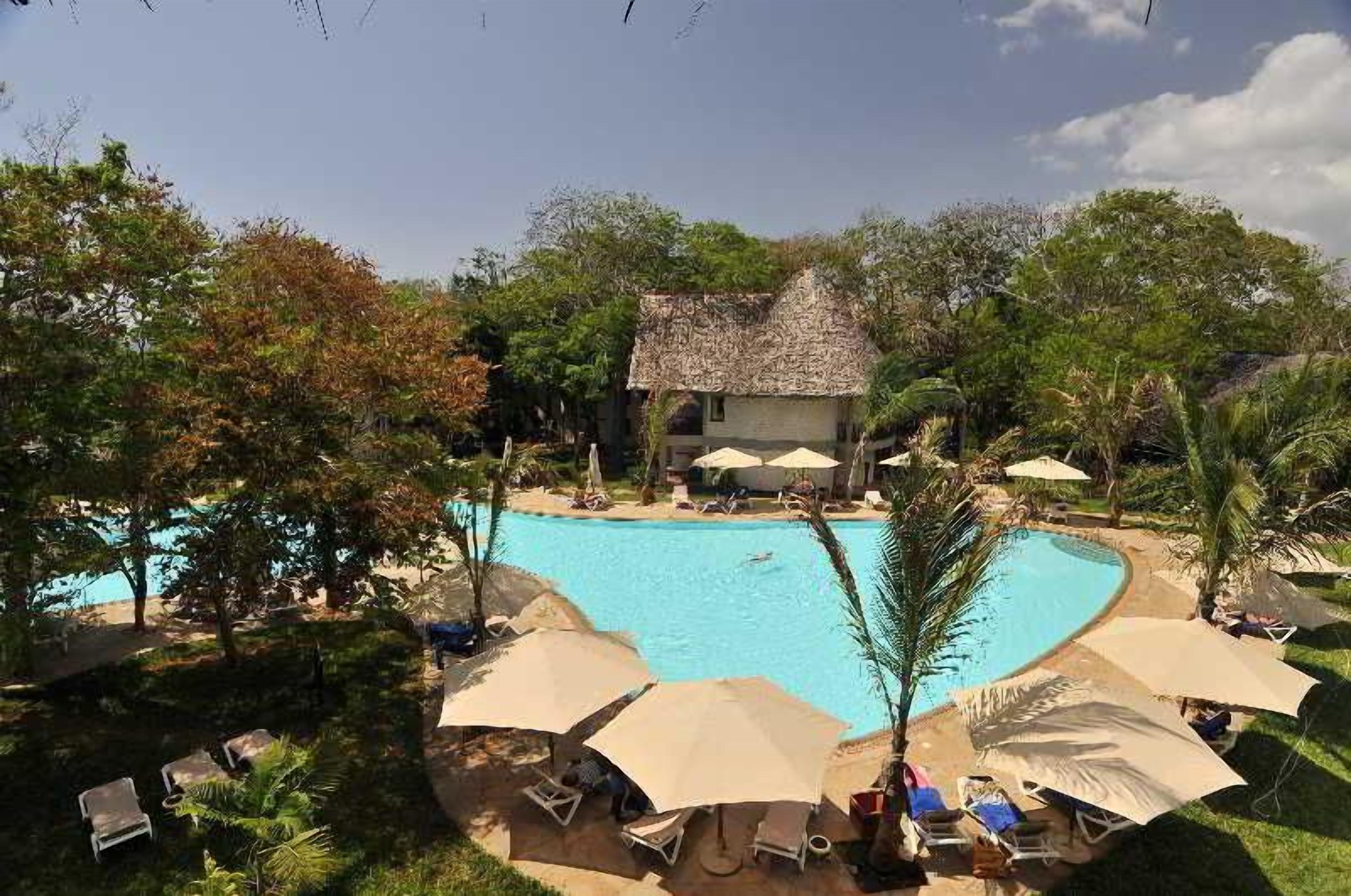 A swimming pool surrounded by lounge chairs and umbrellas, with a thatched-roof building in the background.